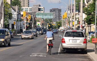 A shared-lane marking in Toronto, Ontario, Canada. Note that the cyclist is not properly positioned on the roadway; they are in the door zone of the parked car and vulnerable to being "doored". Cyclists should ride over the sharrow.