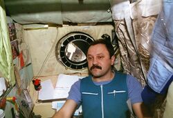 A man, dressed in blue work clothing, seen in a small cubicle. On the walls around him can be seen a sleeping bag, children's drawings, technical manuals and stained insulation. A small porthole in the centre of the wall behind him shows the nose of the Space Shuttle Atlantis and the blackness of space.