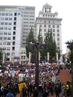A tall, thin structure. In the background are tall buildings. At its base, it is surrounded by crowds of people.