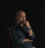 A photograph of a bald man facing right and looking right while covering his mouth with his left hand and wearing a dress shirt all on a black background