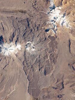 A volcano surrounded by expanding lava flows, left and right two snow covered summits, as seen from space
