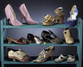 Three blue shelves on a black background. The top shelf displays a pair of pink high heels, a wedge sandal and a dance heel shoe. The middle shelf displays a variety of low-heeled, slipper-type shoes. The bottom shelf displays two antique heeled shoes and a pair of kid's shoes.