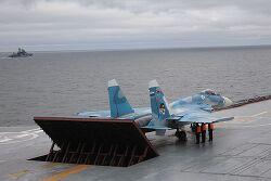 Rear port view of aqua-and-white jet aircraft lining up on aircraft carrier's deck, preparing for takeoff. The jet blast deflector is erected behind the aircraft. Three men in bright orange fluorescent tops stand underneath the jet's right wing