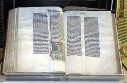 A large old Bible, open and displaying two pages of densely-written calligraphy, with some decoration surrounding one section on the left-hand page.