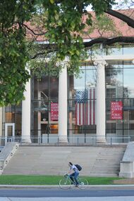 A photo of the John Glenn College of Public Affairs, with an American flag hanging inside and a cyclist riding past the stone steps