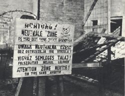 A multilingual sign (in German, Polish, Hungarian and French) warning people against trespassing for fear of being shot. Broken fence with some barbed wire appears behind the sign