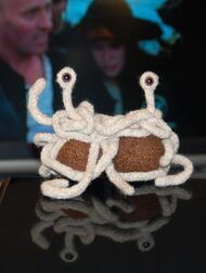 small handmade knit Flying Spaghetti Monster sitting on a table with people dressed as pirates in background.