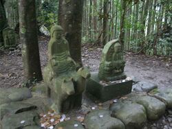 An ancient statue of a young boy riding a cow and a horse at the same time by straddling both at once. The boy is an early emperor on the Kumano pilgrimage. It is set within the forest and is near sacred Buddhist sites.