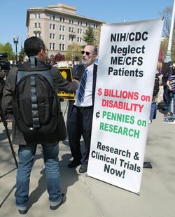 A man with a large sign that reads "NIH/CDC Neglect ME/CFS Patients. BILLIONS on DISABILITY, PENNIES on RESEARCH. Research & Clinical Trials Now!"