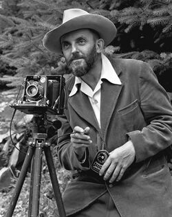A photo of a bearded Ansel Adams with a camera on a tripod and a light meter in his hand. Adams is wearing a dark jacket and a white shirt, and the open shirt collar is spread over the lapel of his jacket. He is holding a cable release for the camera, and there is a rocky hillside behind him. The photo was taken by J. Malcolm Greany and first appeared in the 1950 Yosemite Field School Yearbook.