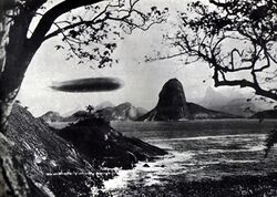 A black-and-white photograph taken between two trees. The Graf Zeppelin is flying at low level from left to right above a rocky coastline. In the background is the distinctive point known as Sugar Loaf, which marks the entrance to Rio de Janeiro harbour.