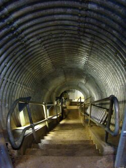 Downward tunnel with steps descending within, flanked by metal handrails. The tunnel is circular in cross-section and lined at the sides and above with corrugated steel.