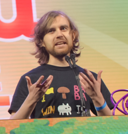 Arvi Teikari speaking behind a podium. He is wearing a black shirt with many icons from the game, including a flag, a mushroom, the "BABA", "IS", and "WIN" operators, and Baba itself.