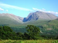 A picture of Ben Nevis on a clear day. A clear blue sky, a large mountain and a very green forest.