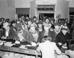 A large crowd of sullen looking workmen at a counter where two women are writing. Some of the workmen are wearing identify photographs of themselves on their hats.