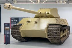 A frontal view of a large, pale-yellow tank in a white museum gallery.. Its curved-faced turret is pointing forwards, the long gun overhangs the front by several meters.