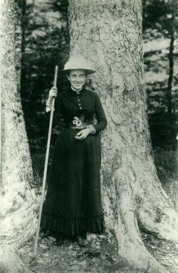 A white woman smiling and standing in front of a tree trunk. She is wearing a dark dress and a wide-brimmed hat and is holding a walking stick.
