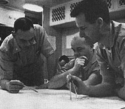 Three naval officer plotting the submerged course of the nuclear submarine Triton, with Captain Beach seated before chart in the center, with Lt. Commander Will M. Adams standing at right holding a pair of dividers and Lt. Commander Robert W. Bulmer standing at the left holding a pencil, and an unidentified individual seated in the background.