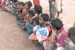 Row of young children sitting or squatting in the dirt wearing dirty clothing, smeared in excrement, one child clutches two towels