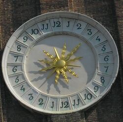 Decorative clay/stone circular off-white sundial with bright gold stylized sunburst in center of the 24-hour clock face, one through twelve clockwise on right, and one through twelve again clockwise on left, with J shapes where ones' digits would be expected when numbering the clock hours. Shadow suggests 3 PM toward the lower left.