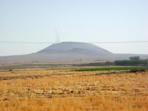 A black mountain with a telecommunication tower above it, seen from afar, and distorted by dusty mist. You are standing in a field overgrown by dry grasses, some of them are green.