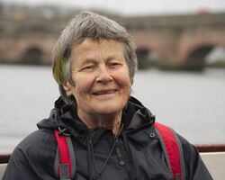 A photo of Dinah smiling, wearing a waterproof jacket, in Berwick on Tweed. A section of her hair, which is otherwise grey, has been dyed orange, yellow and green.