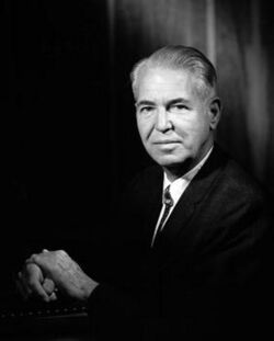 Upper body of a serious and dignified man in his fifties, with graying hair combed back and in a dress jacket with white shirt and a bolo tie and with his hands coupled together on top of some books.