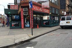 View of the triangle, which is located on a sidewalk at a street corner. The triangle is outside the Village Cigars shop and the Christopher Street–Sheridan Square station of the New York City Subway. The triangle can be seen on the sidewalk toward the left side of the photo.