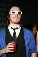 Fish wears his medium-long hair off his face, some stubble facial hair, 3D glasses, a white dress shirt with loosened black tie under a black vest and a blue cardigan while holding a red drink at a party.