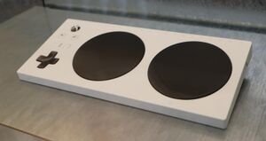 Xbox Adaptive controller, which is a large white rectangular slab with a few basic controls, mostly for Xbox console system functions, and multiple ports for accessible input devices. The controller itself consists of a white plastic slab with a slight slope from back to front; the right side of the controller is taken up by two large black dome buttons which default to controller inputs A and B; the left side of the controller has (from back to front) an Xbox system button, the View and Menu buttons, a button to switch between one of three saved mapping profiles, and a cross or plus-shaped digital directional pad.