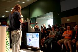 A man wearing a black shirt and jeans and carrying a microphone speaks to a sitting crowd. In the center of the frame is a flatscreen television displaying Halo Wars gameplay.