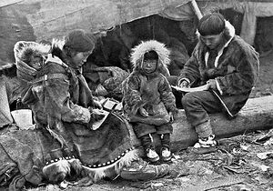 An Inuit family is sitting on a log outside their tent. The parents, wearing warm clothing made of animal skins, are engaged in domestic tasks. Between them sits a toddler, also in skin clothes, staring at the camera. On the mother's back is a baby in a papoose.