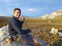 Image of Sean Dougherty in climbing gear. He is sitting on a rock on the left side of the image, smiling to the camera, while wearing technical climbing gear and handling a climbing rope.