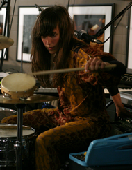 A 32-year-old woman with long brown hair, and a yellow and brown outfit. She is playing drums, and is surrounded by music equipment.