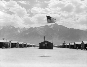 Small buildings in rows in a dusty flat area. A tattered U.S. flag flies from a pole in the foreground, and tall mountains dominate the background below a sky with clouds.