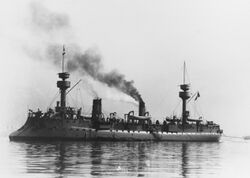 A large gray ship sits at anchor, riding high in the water. Thick black smoke drifts lazily up from two smoke stacks.