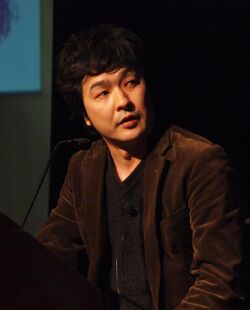 A Japanese man in a brown jacket and dark grey shirt standing at a podium.