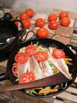 Filets of fish on a bed of shredded vegetables cooking in a frying pan, partially covered by sliced tomatoes.