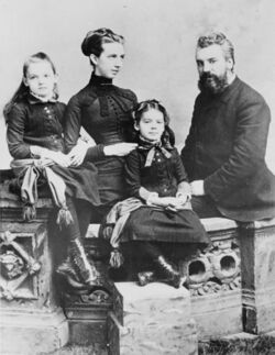 A distinguished bearded man, his young elegant wife next to him and their two young daughters poise for a formal portrait