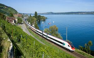 Red, white, and black trains pass a blue lake at speed