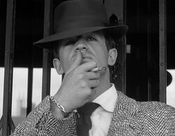 Black-and-white image of a man seen from mid-chest up, wearing a fedora and a jacket with a houndstooth-like pattern. He holds a cigarette between the middle and index fingers of his left hand and strokes his upper lip with his thumb. He stands in front of what appears to be a mirrored doorway.