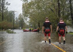 A photograph of two men walking on a flooded street towards a group of men in rafts in the background
