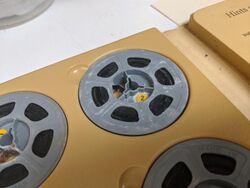 Close up of a film reel of 8mm home movie in an album. The grey plastic reel is covered in a white powdery substance that has spread to the film.