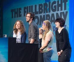 Four smiling people (two women and two men) stand in front of a podium. Behind them is a large blue screen with the words THE FULLBRIGHT COMPANY projected across it.