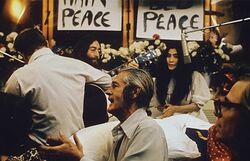 Lennon and Ono sit in front of flowers and placards bearing the word "peace". Lennon is only partly visible, and he holds an acoustic guitar. Ono wears a white dress, and there is a hanging microphone in front of her. In the foreground of the image are three men, one of them a guitarist facing away, and a woman.