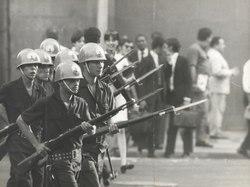 Several men in a line wear helmets and brandish rifles with bayonets