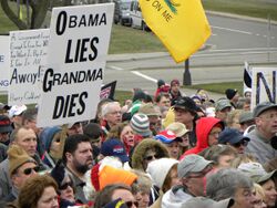 A group of people near a street corner in the background, seen from above head level. At the left one holds up a black-on-white sign saying "Obama Lies, Grandma Dies". Portions of other signs are visible and part of a Gadsden flag hangs from above the top of the image