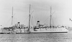 A light gray ship with two widely spaced funnels and three masts lies at anchor with small boats alongside.