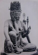 Three-quarter view of a seated statue with six arms. The left leg is bent, touching the ground along its whole length. The right leg is also bent, but with the knee up. The right foot is above the left foot. One of the right hands is touching the right ear, another is lifting a jewel-shaped object in front of the breast. One of the left hands carries a flower, another a small wheel-shaped object.