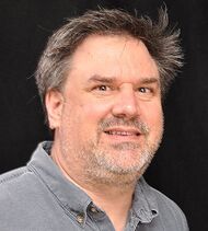 A brown haired man in a gray shirt looks into the camera while standing against black background.